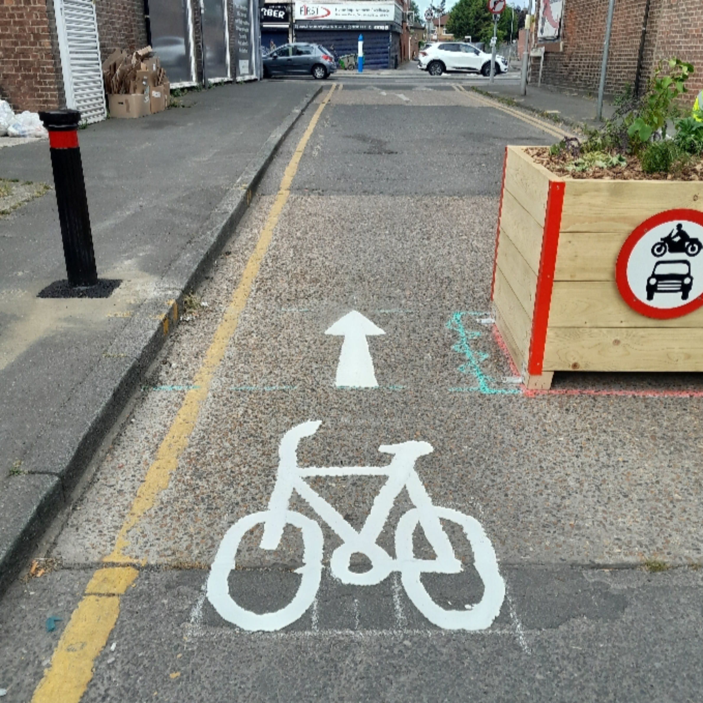 A painted white bicycle symbol and straight arrow on a asphalt road lane, leading past a wooden planter box with a red "no motor vehicles" sign on a urban street.